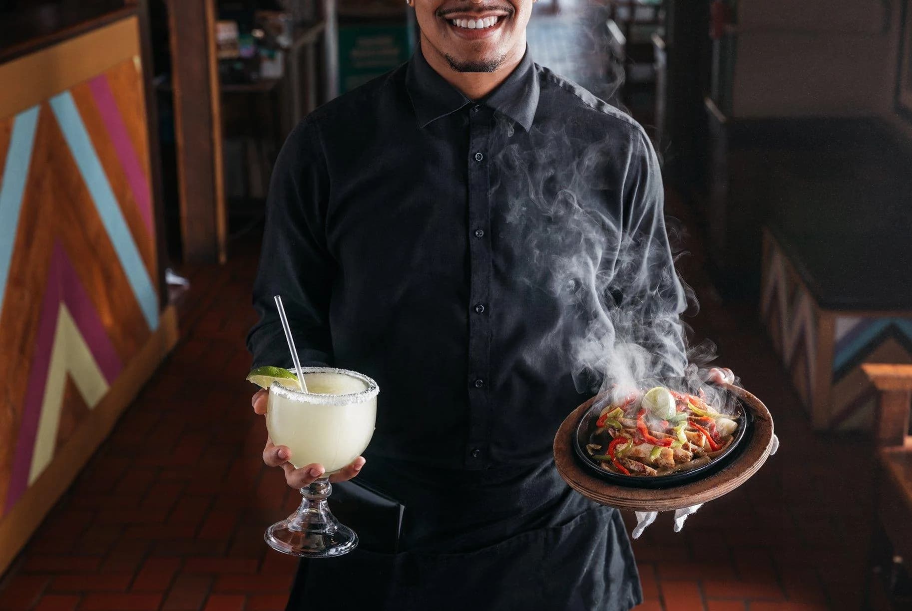 Smiling server carrying a frozen margarita and a sizzling plate of fajitas